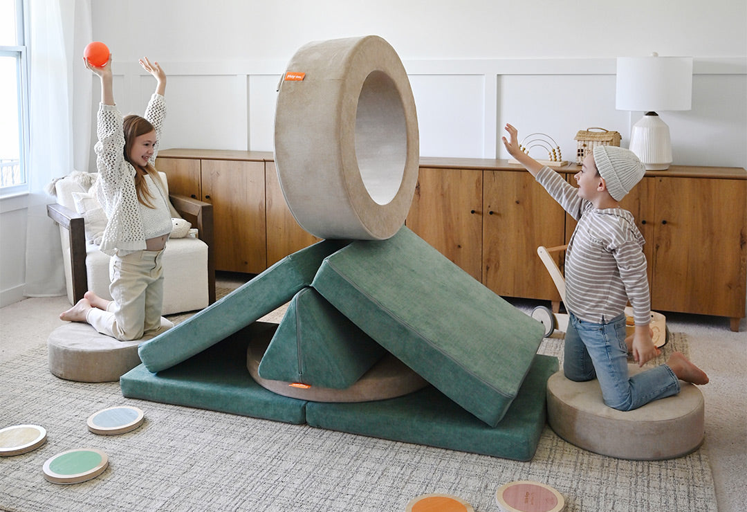 Two children smile and cheer as they play a newly-invented game with a sea green Nugget and a beige play ottoman.