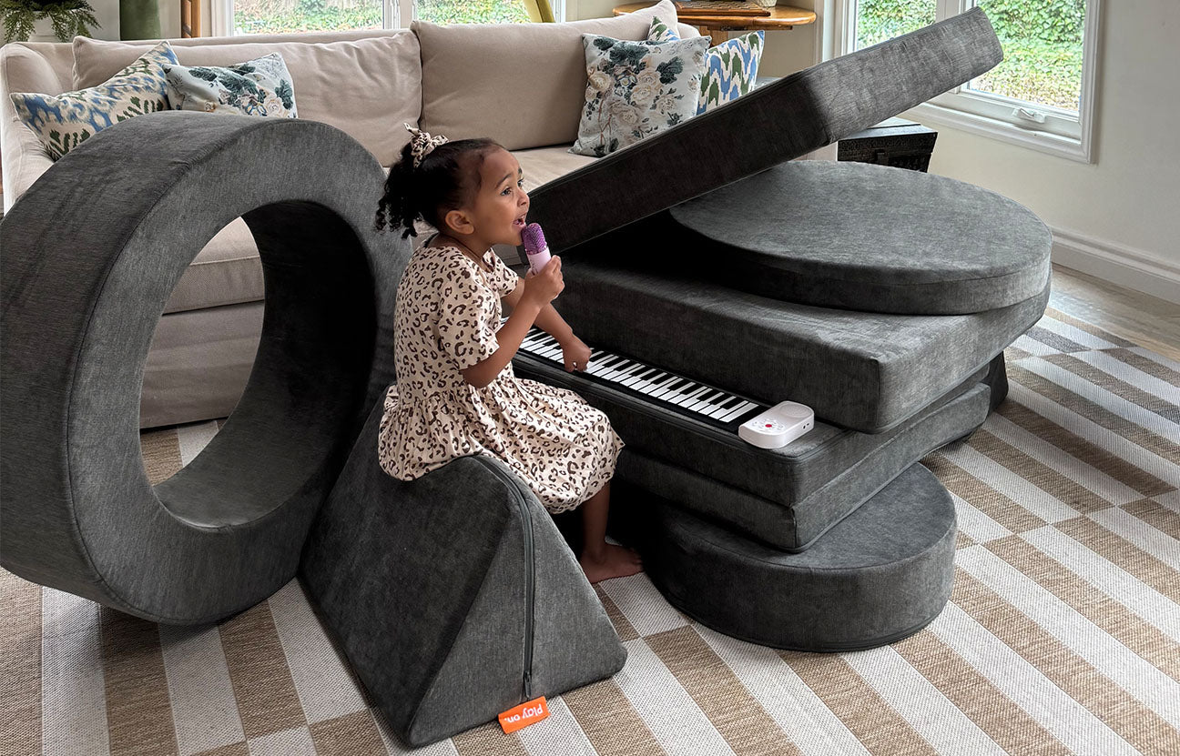 A child sings and plays a pretend baby grand piano built out of a black Nugget play couch and Chunk play ottoman.