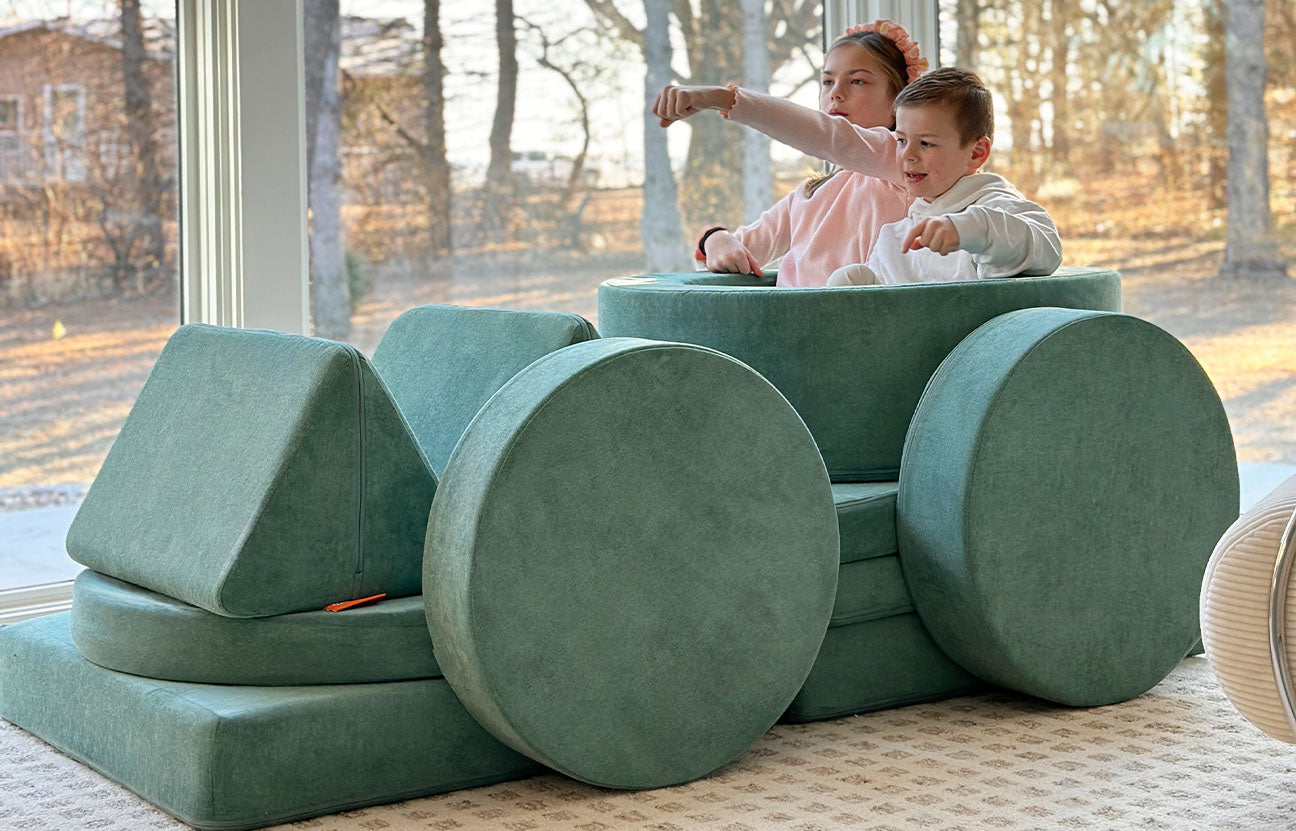 Two children pretend to drive a racecar built out of a sea green Nugget play couch and Chunk play ottoman.