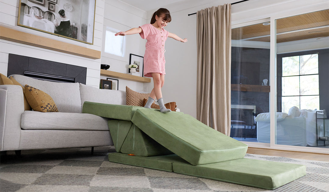 A child balances on a Bamboo mossy sage Nugget play couch, set up as a ramp onto the conventional family couch.