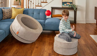 A child throws a ball over his shoulder into the hoop of a Dunebuggy vintage beige Chunk play ottoman.