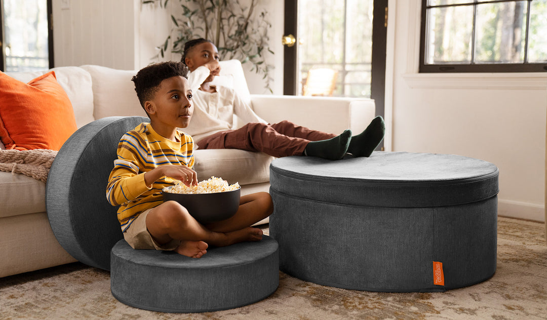 A child sits on a puck of a Harbor charcoal gray Chunk play ottoman, while a second child props his feet on the hoop and pad set up as an ottoman.