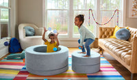 Two smiling children play on a Surfside light gray-blue Chunk play ottoman, set up as a ball pit and platform.