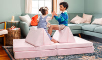 Two children sit on triangular pillows and high five each other, atop the flat pieces of a Heartthrob blush pink play couch by Nugget.