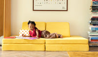 A young child reads a book on a Lemonade classic yellow play couch by Nugget.