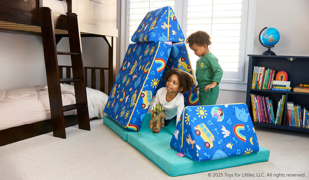 Two children play on the pieces of a vibrant Ms. Rachel-patterned Nugget play couch, set up as a tent and hurdle.