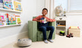 A child seated on a mossy sage green Smore play chair in a bedroom.