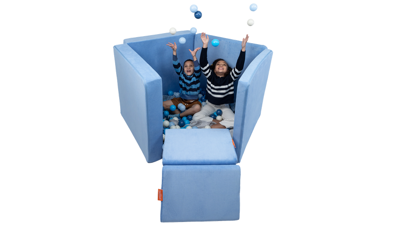 Two children toss balls and play in a Nugget ball pit build, created with a light blue Nugget play couch.