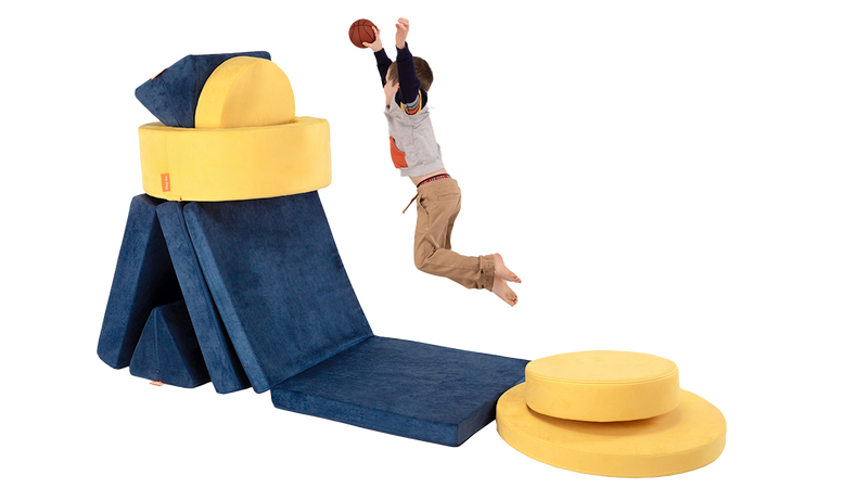 A child dunks a basketball into a hoop made from one navy Nugget play couch and one yellow Chunk play ottoman.