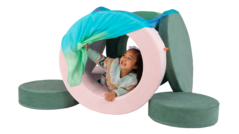 A child plays in a cozy Nugget fort built from two pink and green Chunk play ottomans, plus a Playsilk curtain.