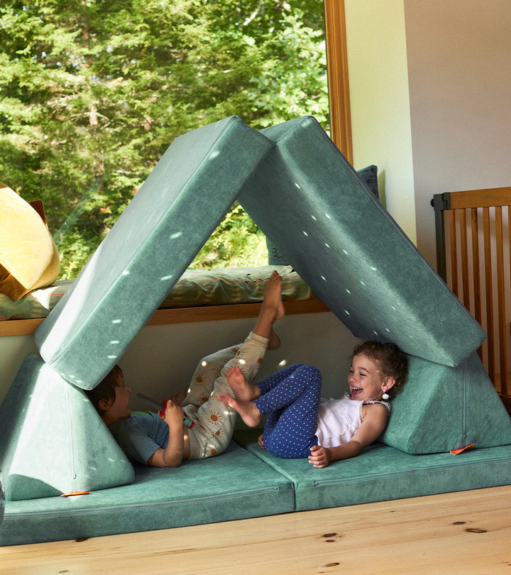 Two children recline and play inside a fort made from a sea green Nugget play couch. 