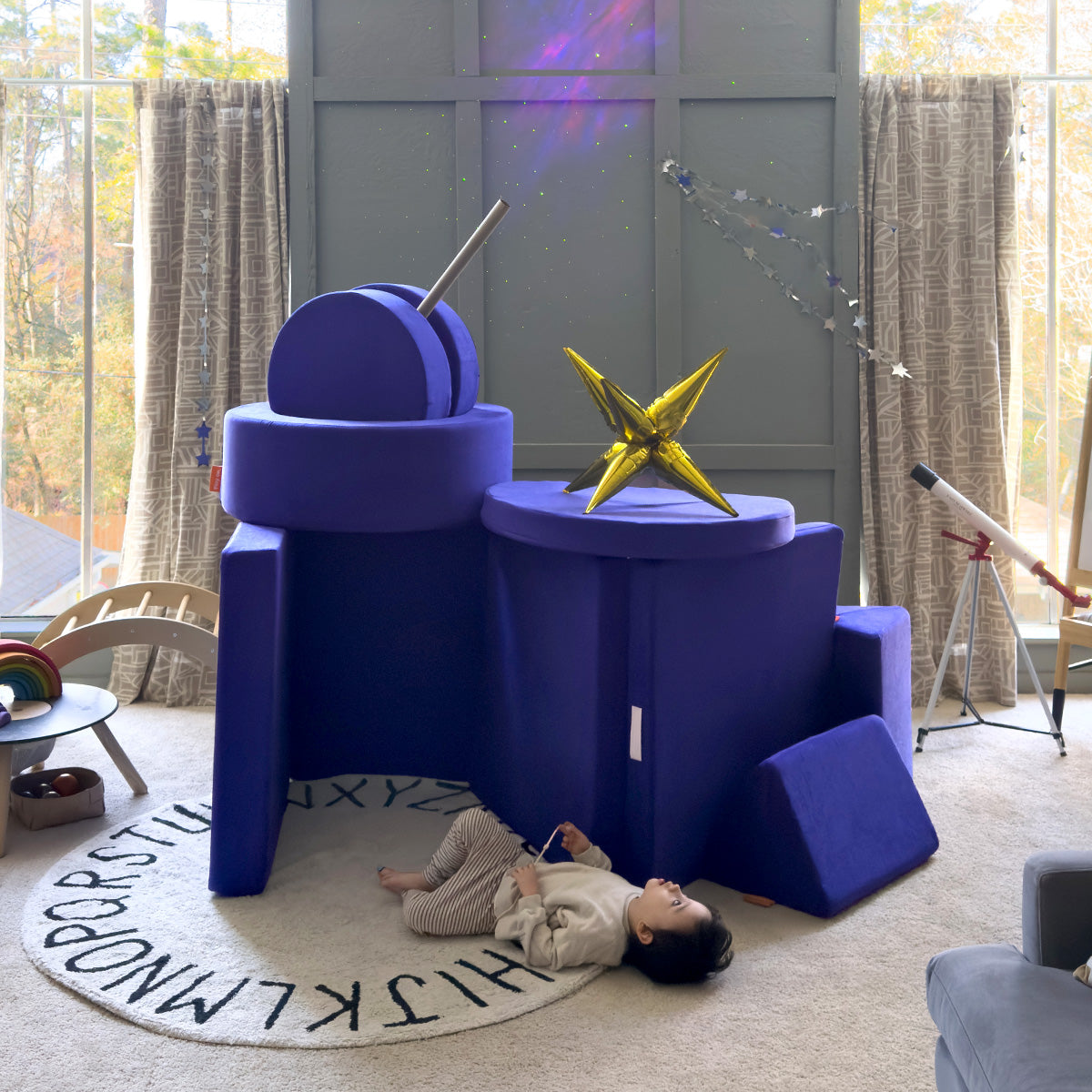 A child pretends to stargaze alongside a pretend observatory built from an indigo Nugget play couch and Chunk play ottoman.