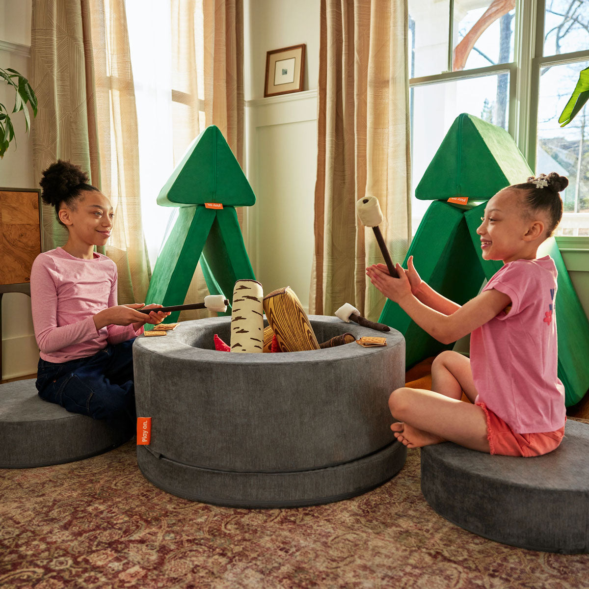 Two kids pretend to roast marshmallows at a campsite built from a green Nugget play couch and gray Chunk play ottoman.