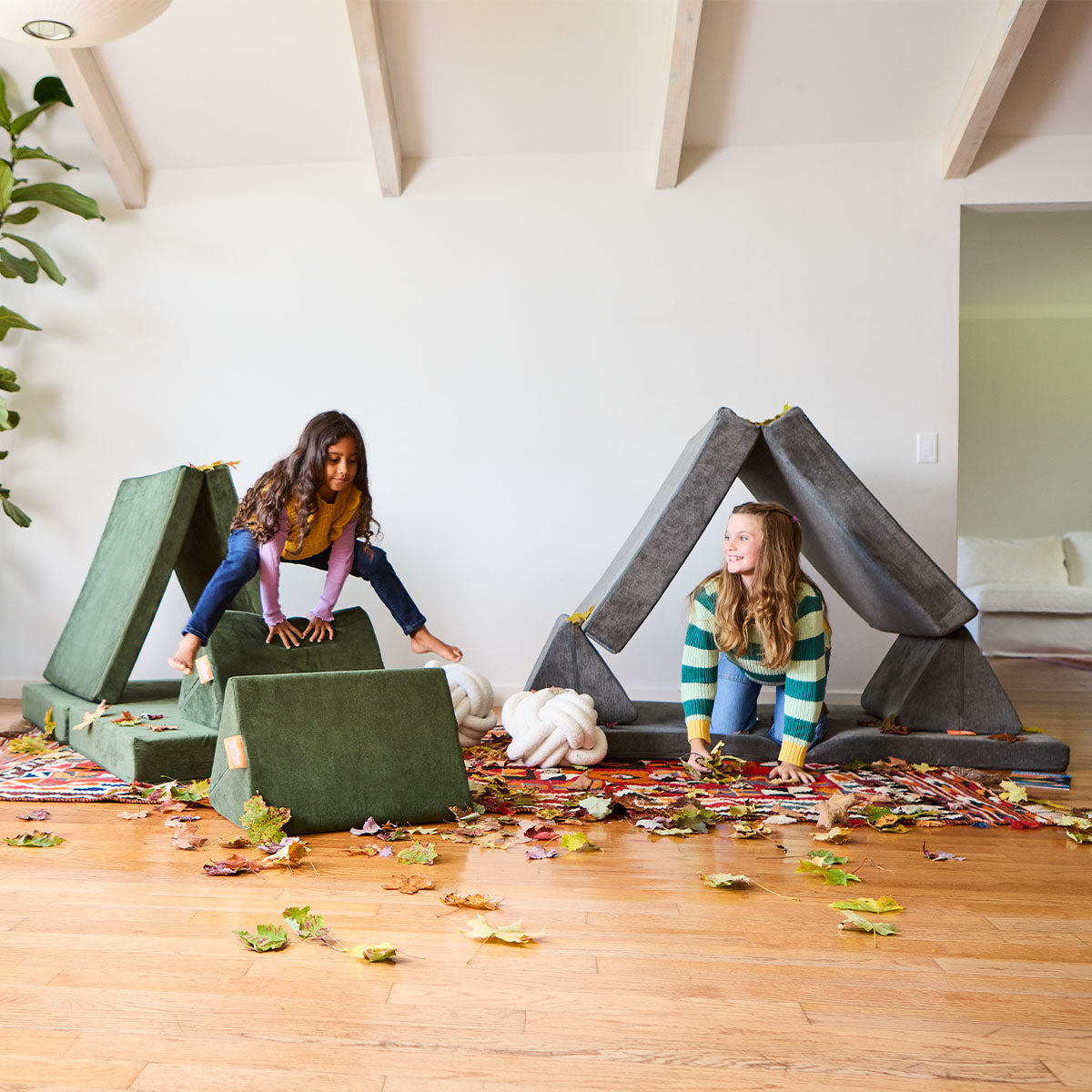 Two children jump and play among fake leaves inside two small structures built from Nugget play couches. 