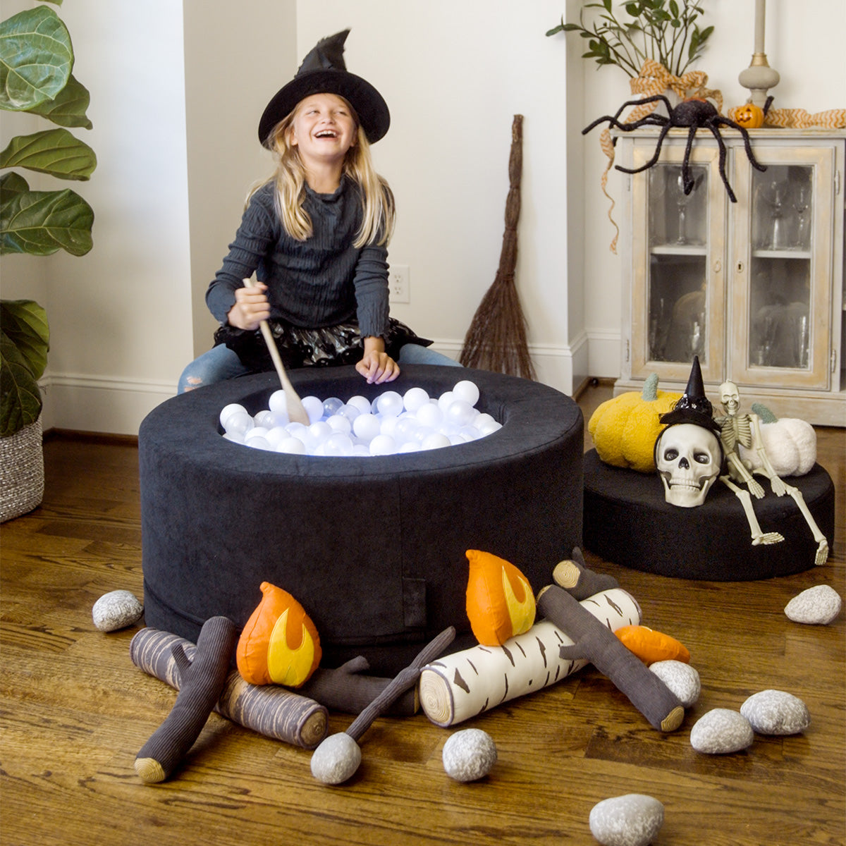 A child dressed as a witch pretends to stir a potion inside a cauldron made from a black Chunk play ottoman.