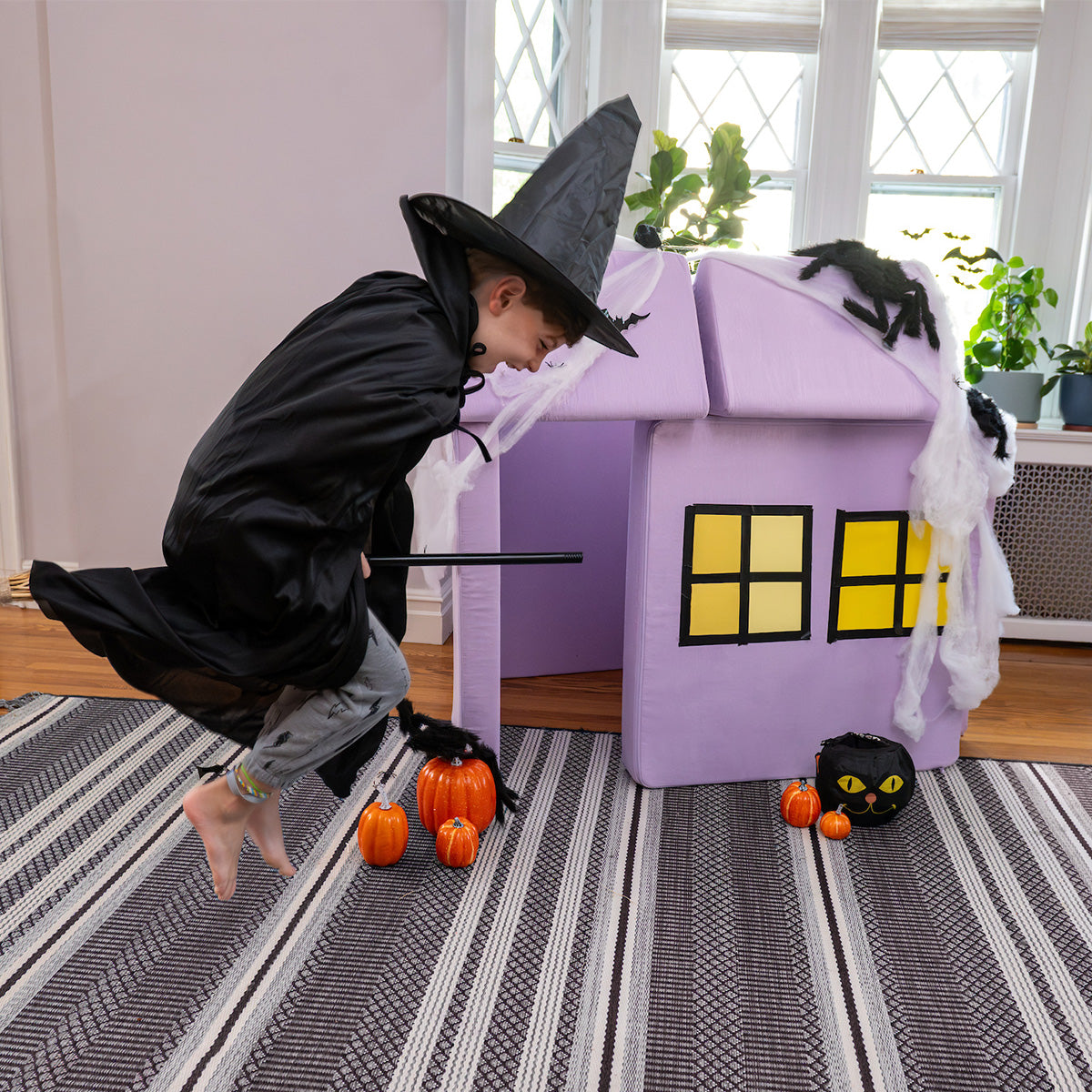 A child pretends to fly on a broom next to a fort built from a lilac purple Nugget.