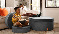 A child sits on a puck of a Harbor charcoal gray Chunk play ottoman, while a second child props his feet on the hoop and pad set up as an ottoman.