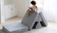 A young child climbs on a Koala light gray play couch by Nugget, set up as a climbing structure.