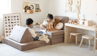 Two children relax and engage in calm play while seated on the cushions of a Kodiak taupe play couch by Nugget using the triangular pieces as backrests.