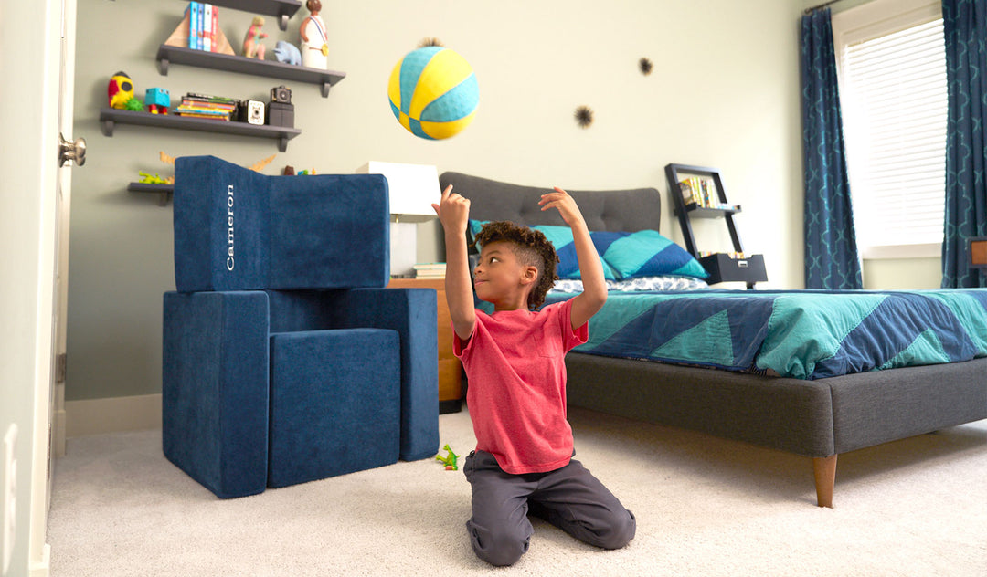 A child tosses a ball backwards into a basket built from the three pieces of a navy blue Smore play chair.