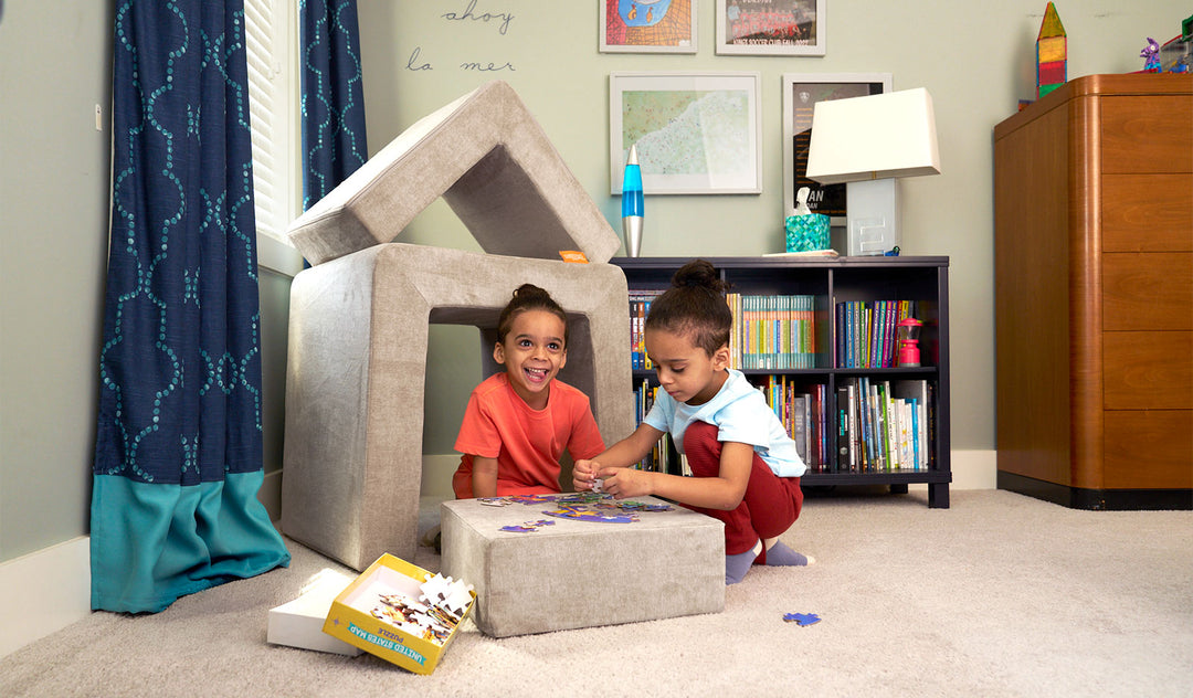 Two children put together a puzzle on the block of a beige Smore play chair, beside a small fort made of the other two Smore pieces.