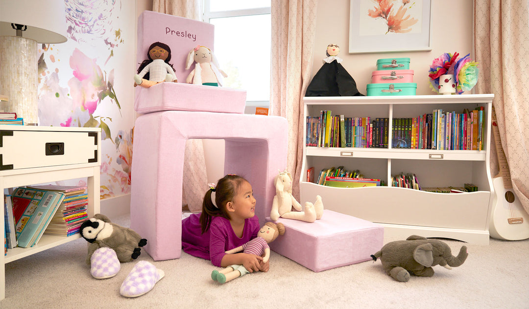 A child plays with dolls and stuffies inside the pieces of a blush pink Smore play chair.
