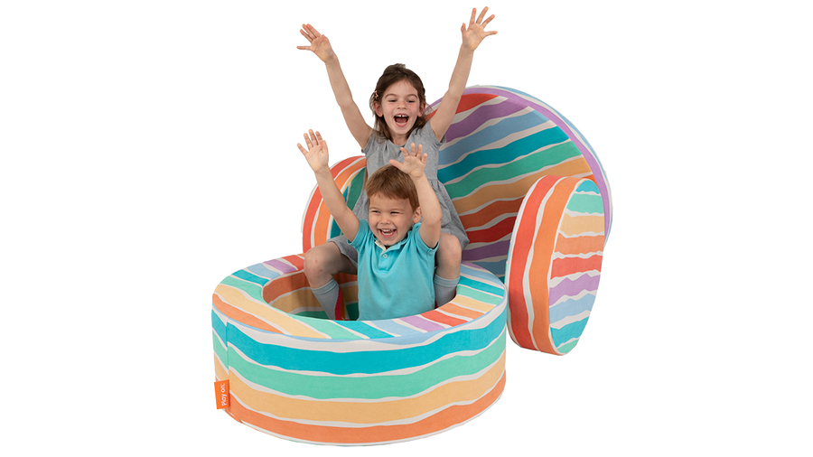 Two children throw their hands in the air as they play inside the hoop of a rainbow-colored Chunk play ottoman.