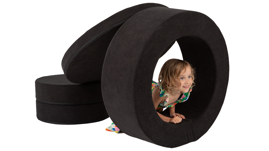A child crouches and peeks through the hoop of a black round storage ottoman, engaging in open-ended play and imagination.
