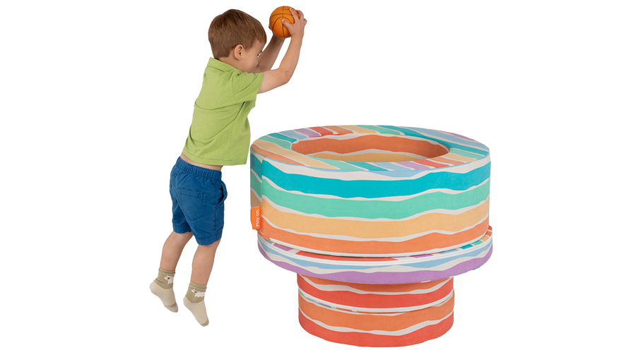 A child leaps in the air and tosses a ball into a basket made from the pieces of a rainbow-colored Chunk play ottoman.