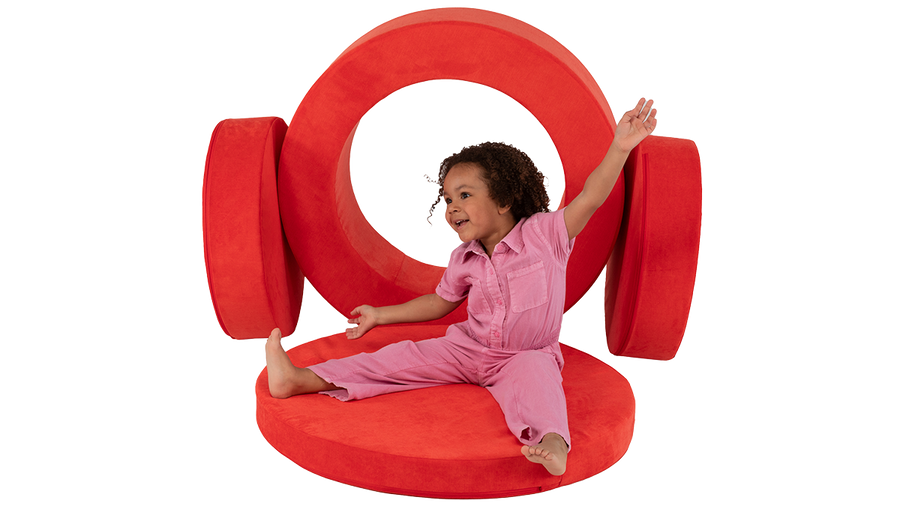 A child poses while playing with the pieces of a red round storage ottoman made by Nugget play furniture.