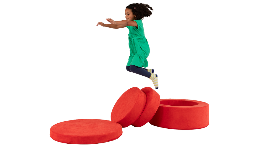 A child is captured mid-air, jumping around on the pieces of a red round storage ottoman called Chunk, made by Nugget.