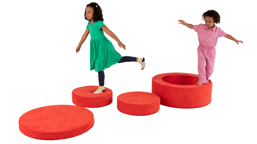 Two children practice their balance on the four foam pieces of a red Chunk play ottoman, engaging in imaginative play.