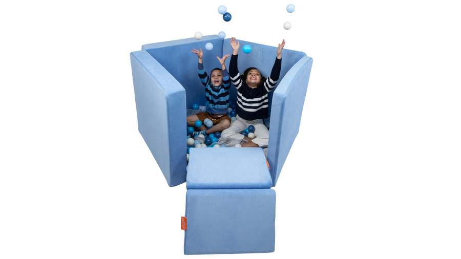 Two children toss balls and play in a Nugget ball pit build, created with a light blue Nugget play couch.