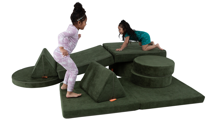 Two children play on a colorful indoor obstacle course built from one Nugget play couch and one Chunk play ottoman. 