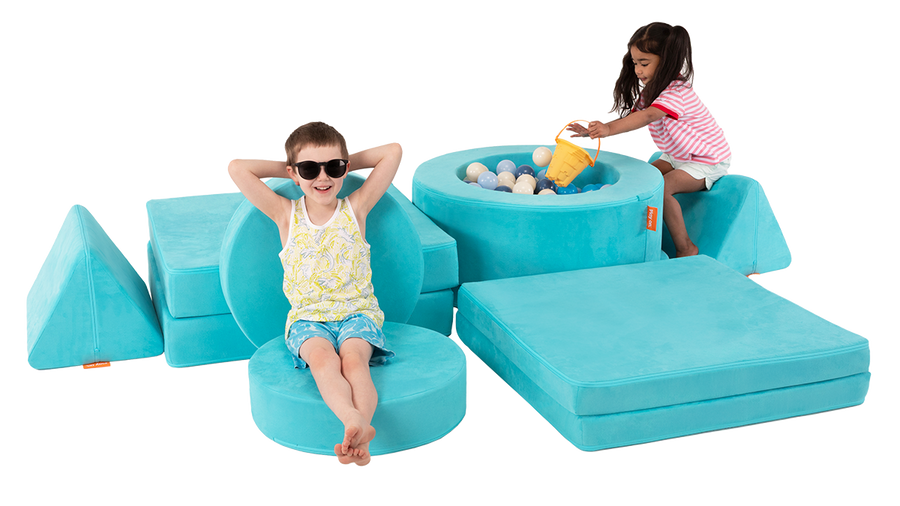 Two kids relax and play on turquoise play furniture — one in a playroom ball pit and the other on a pretend beach chair. 