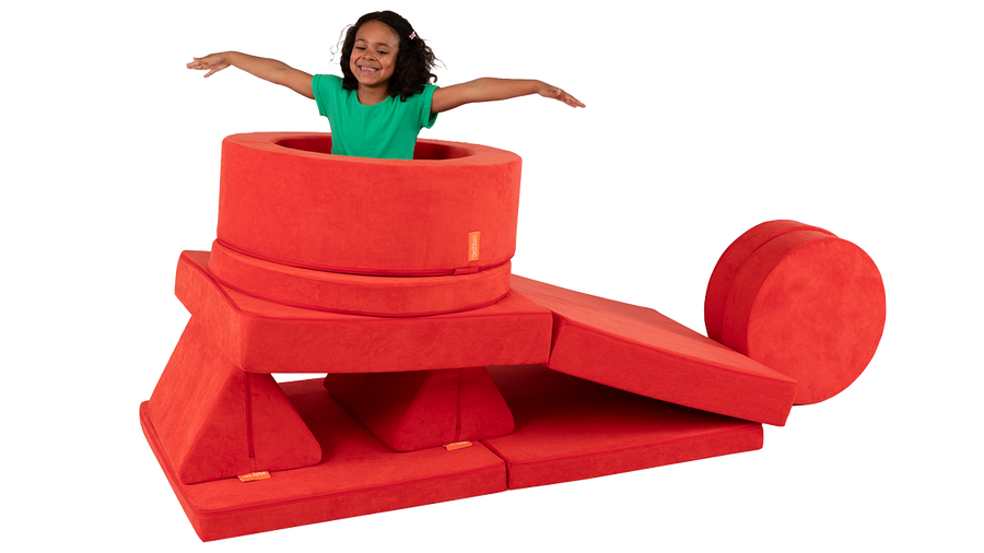 A child smiles and throws her arms out while playing among the pieces of a red Nugget play couch and Chunk storage ottoman.
