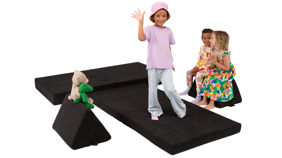 A child struts on a pretend runway made from a black Nugget play couch, with an audience of two friends and several stuffies.