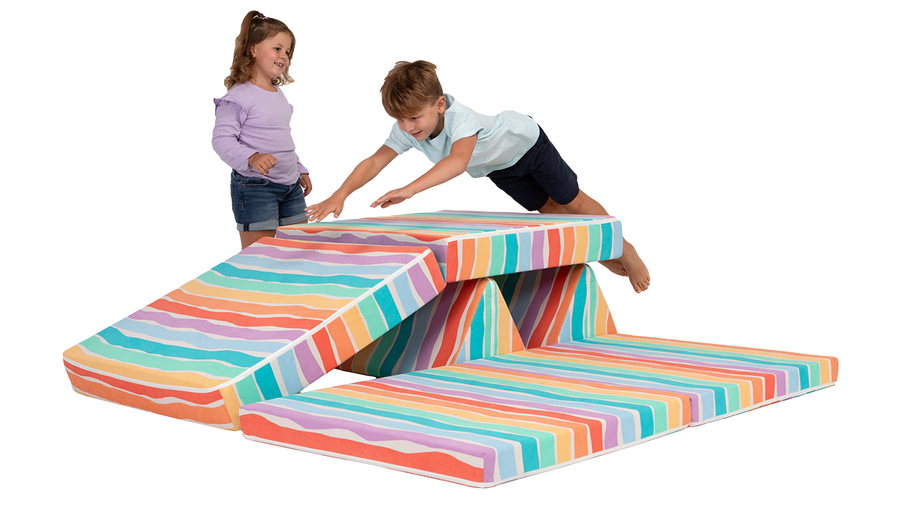 A child jumps across the pieces of a rainbow Nugget play couch, as a second child smiles and watches.