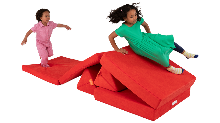 Two children climb and jump on the microsuede pieces of a poppy red Nugget play couch, engaged in imaginative play.