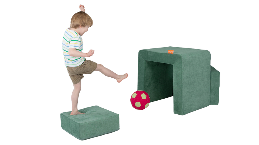A child kicks a ball into the frame of a sea green Smore play chair, set up as a soccer goal.