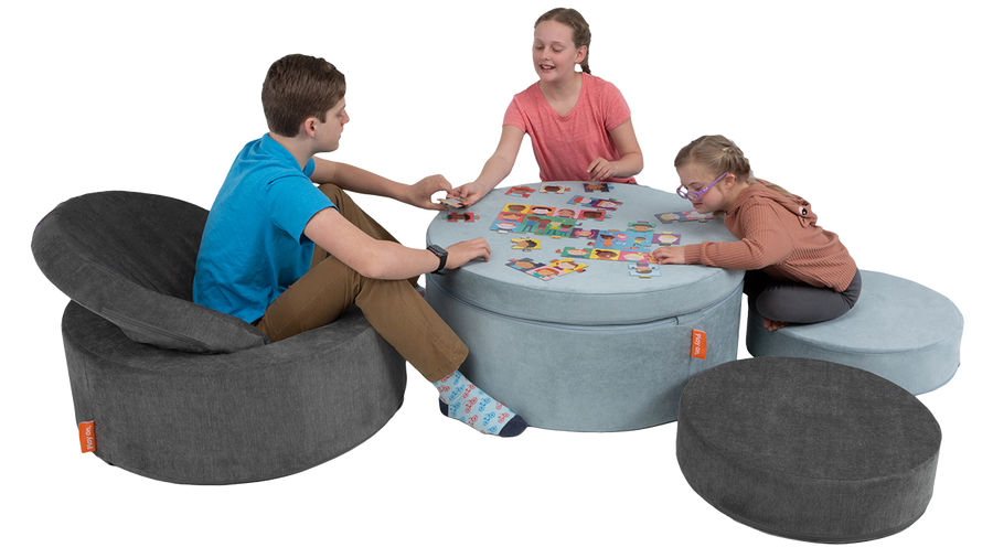 Three children work on a puzzle on a playroom table constructed with two Chunk play ottomans, using the pieces as seating.