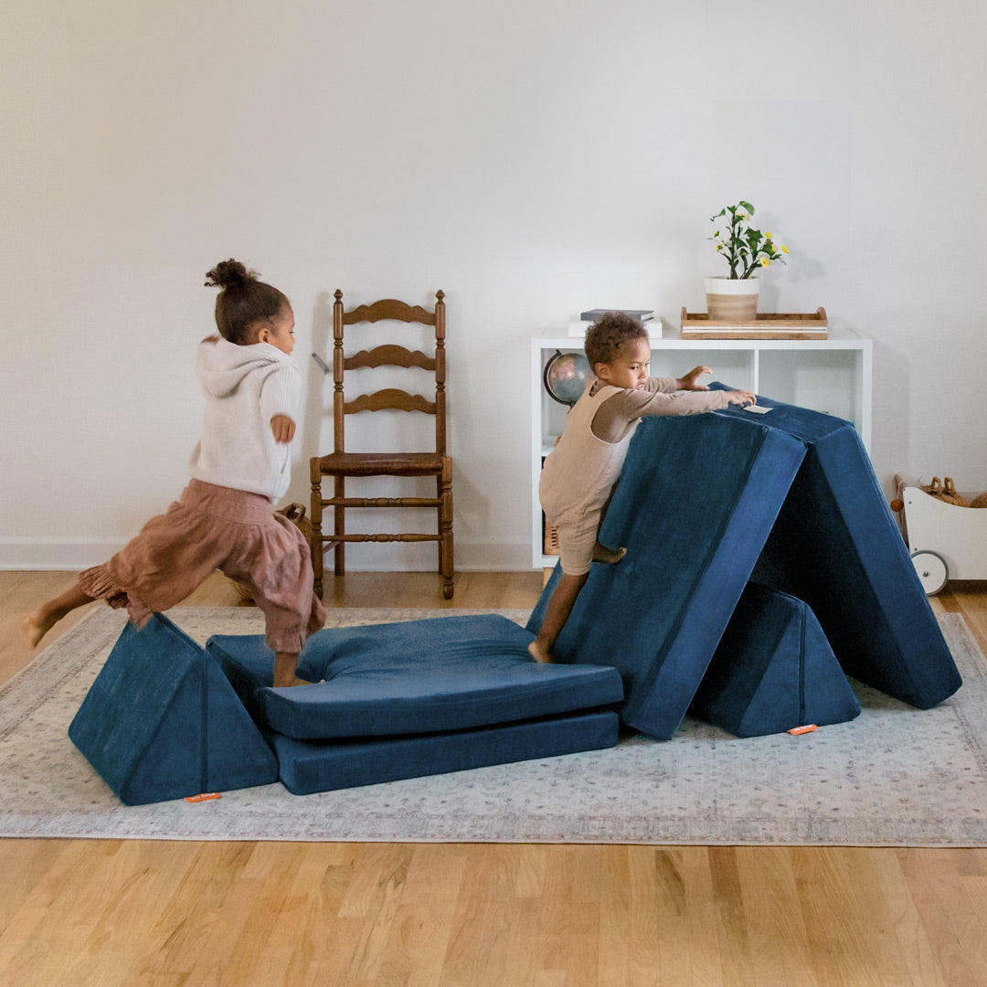 Two children run, jump, and play on a navy blue Nugget® play couch set up to resemble an obstacle course.